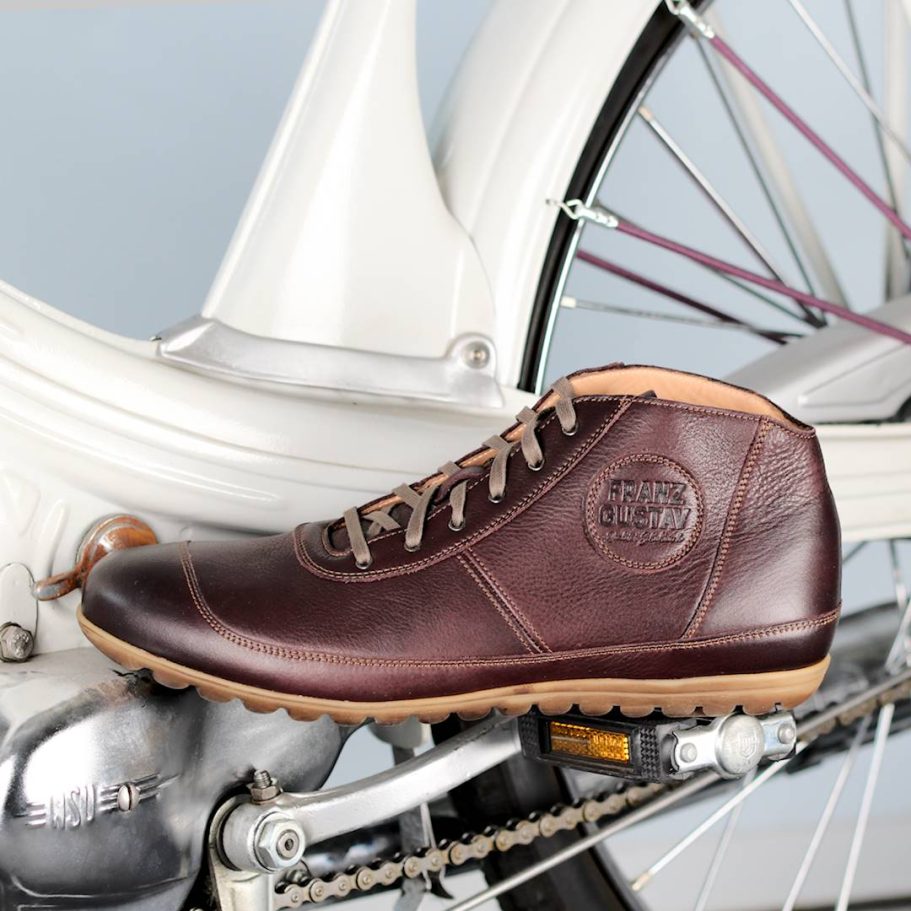 Braune Leder-Schuhe stehen neben einem Fahrrad, auf einem grauen Hintergrund.