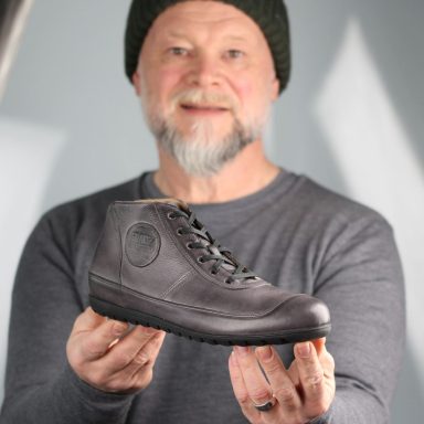 Story Mann hält einen grauen Stiefel in der Hand, lächelnd, mit Mütze und grauem Shirt.